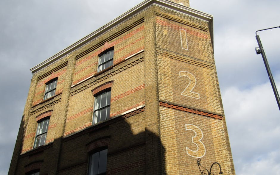 Photograph of a large, multi-story Victorian-style building with red brick façade and ornate white window frames, situated on a city street under a clear blue sky. The building features a prominent r